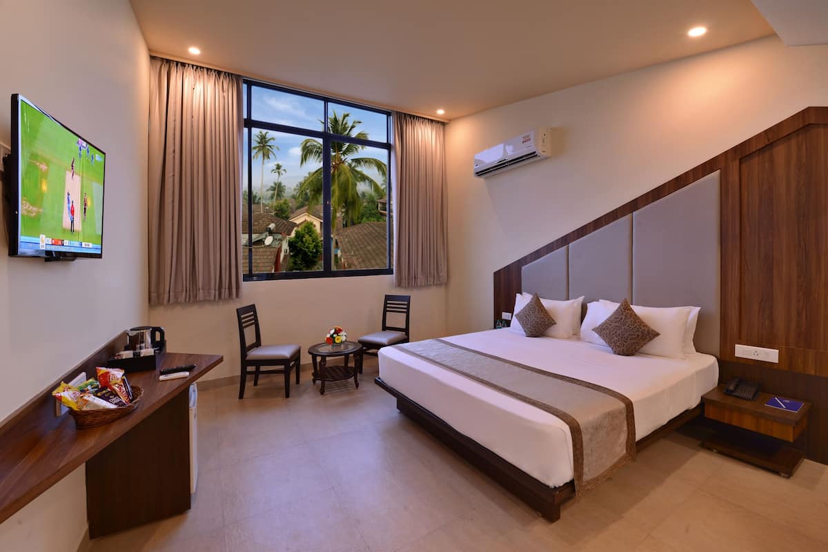 A hotel room with a large bed, a desk with a TV, two chairs, a small table with snacks, and a window with curtains.