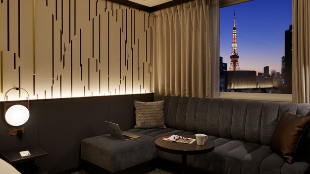Una habitación de hotel moderna con un sofá modular, una mesita redonda pequeña, una computadora portátil y vistas a un paisaje urbano con una torre destacada.