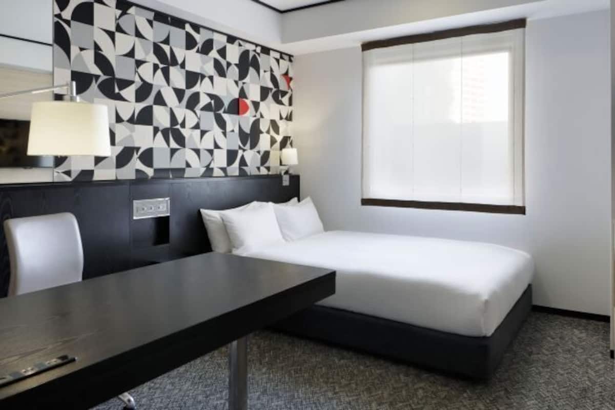 Ett hotellrum med en sÀng, ett skrivbord, en stol, en vÀgg med mönster och ett fönster.