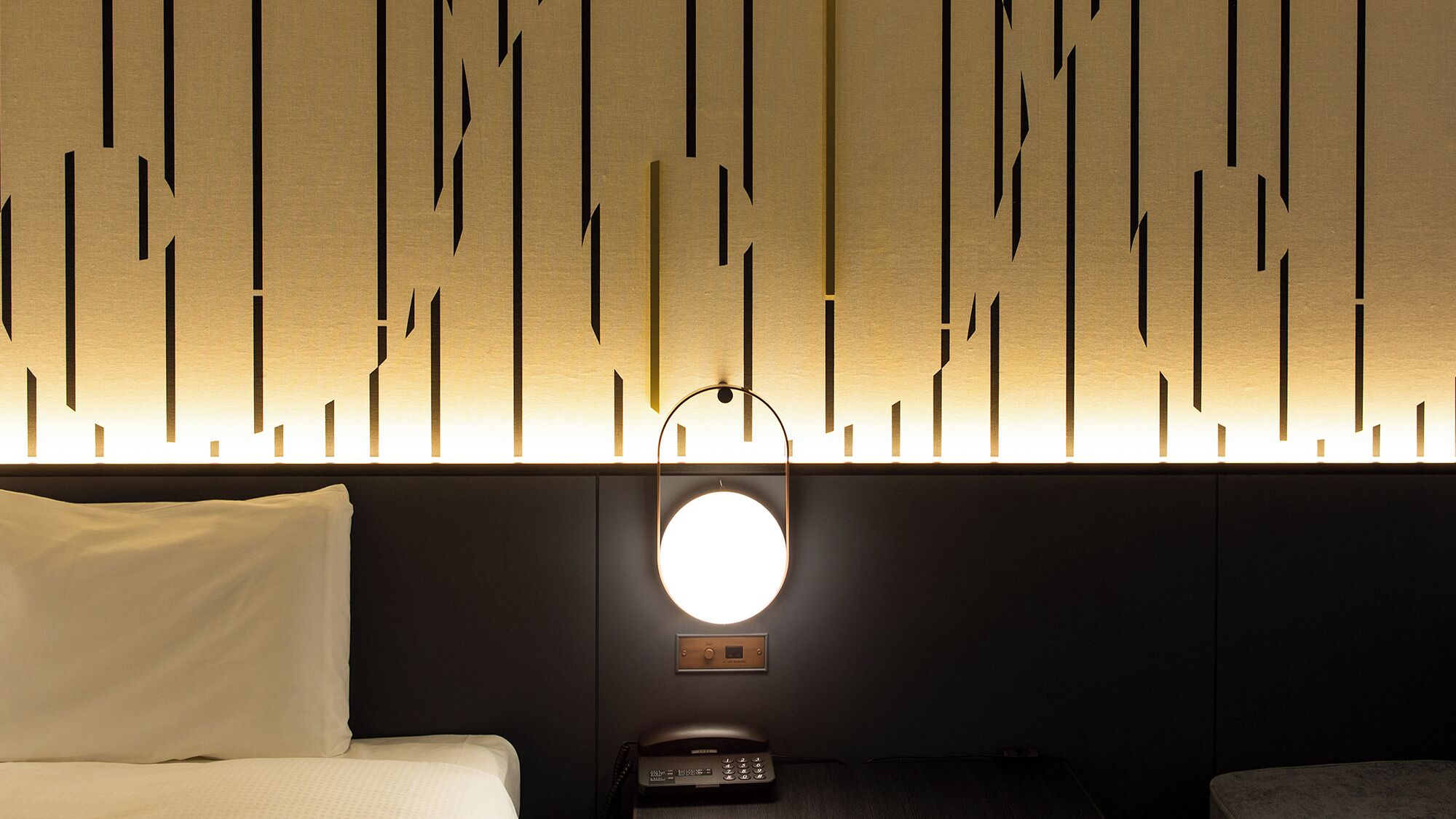 A hotel room with a bed, a bedside table with a phone, and a wall with vertical light fixtures.