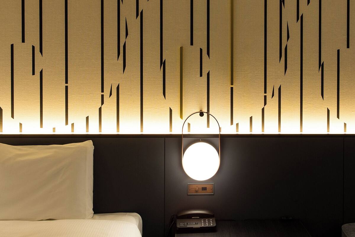 Habitación de hotel con cama, mesita de noche con teléfono y una pared con lámparas verticales.