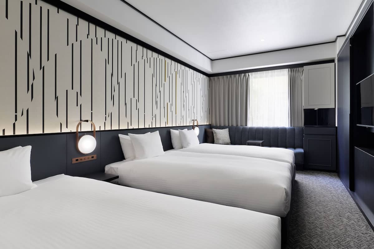 Habitación de hotel con tres camas, un minibar, un ventanal grande con cortinas y un diseño moderno en la pared.
