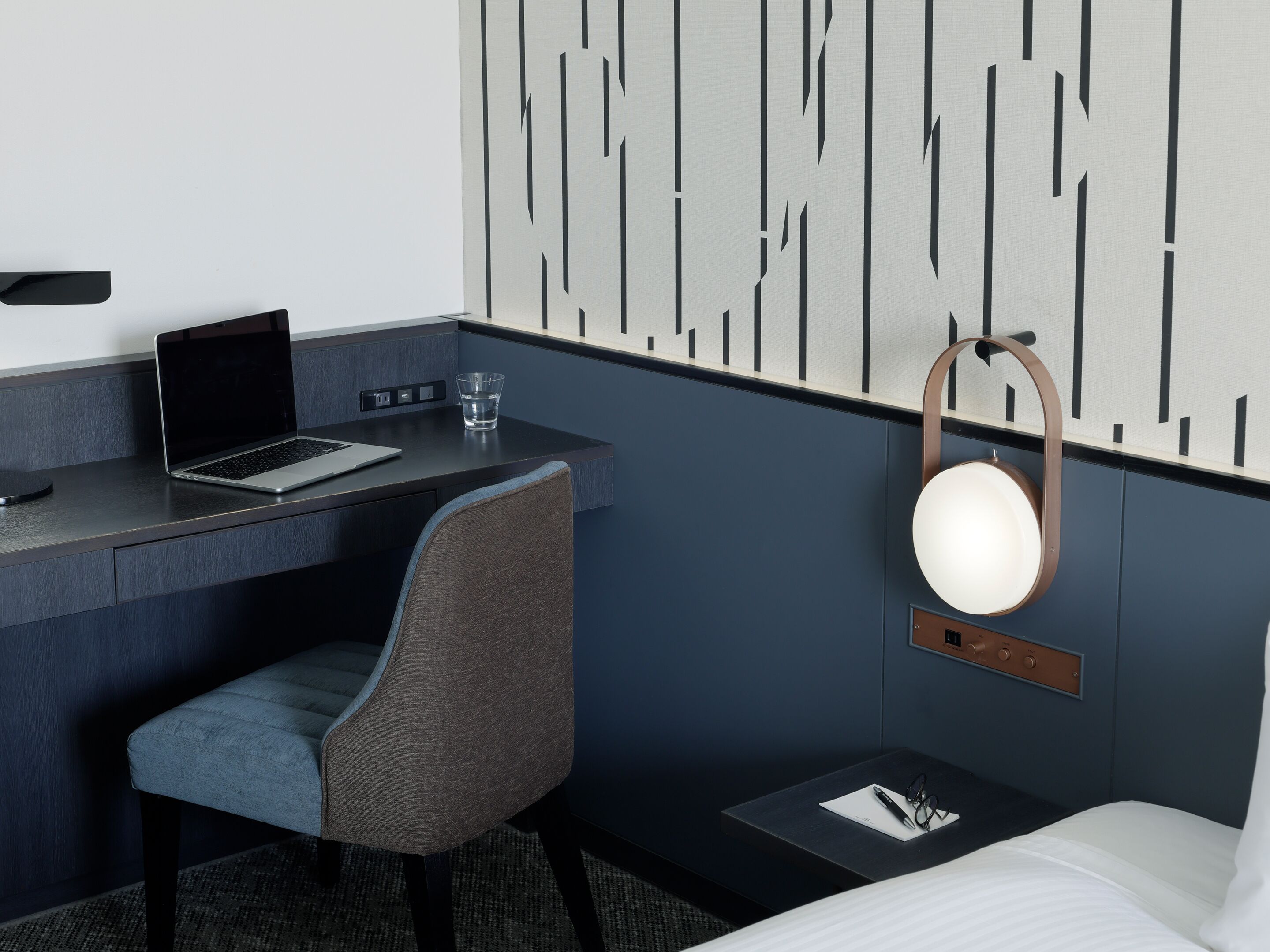 A hotel room with a desk, laptop, chair, bedside table with glasses, and a bed.