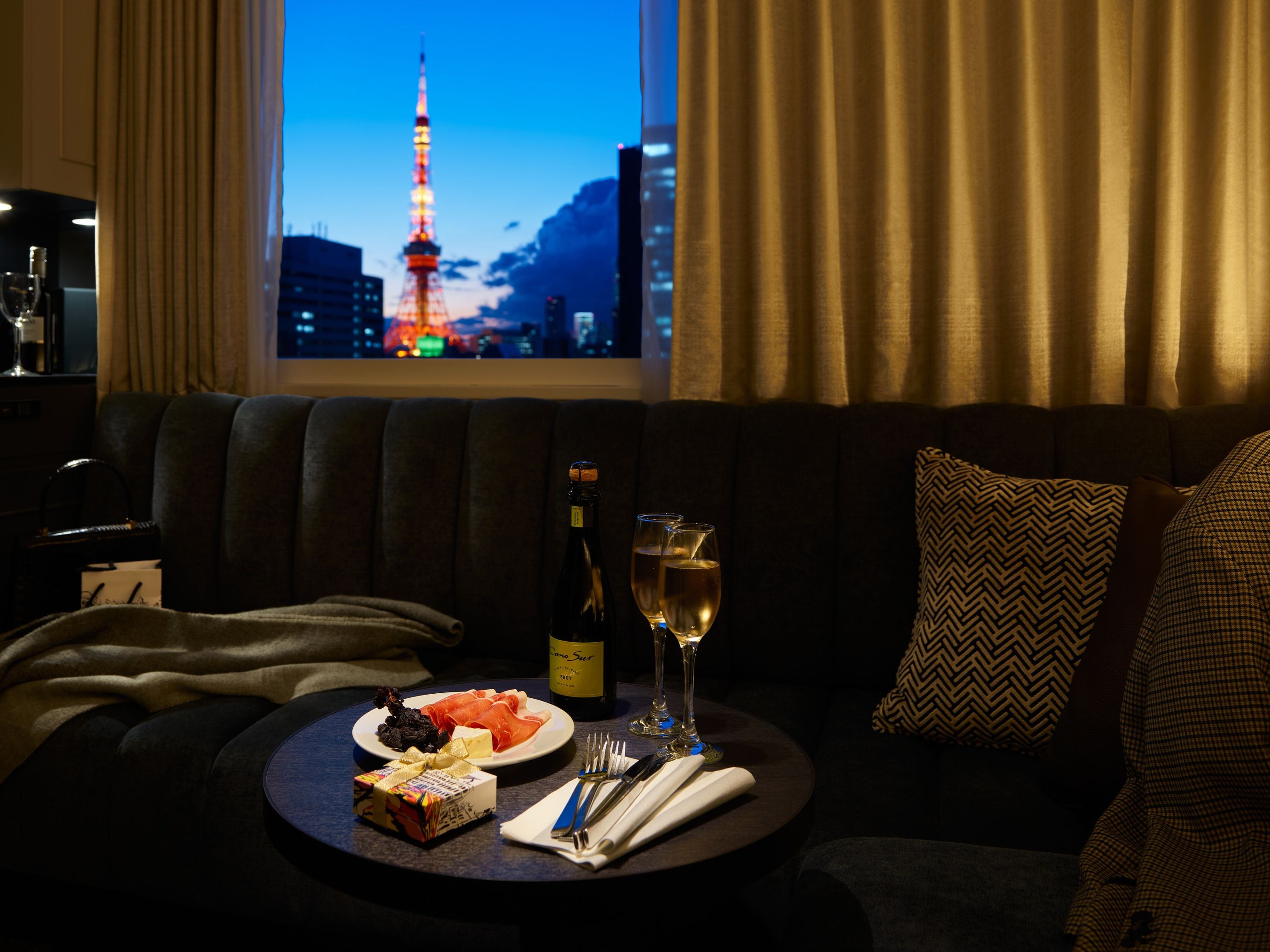 A cozy hotel room with a view of a cityscape, a dining table with a bottle of wine, glasses, and a plate of food.