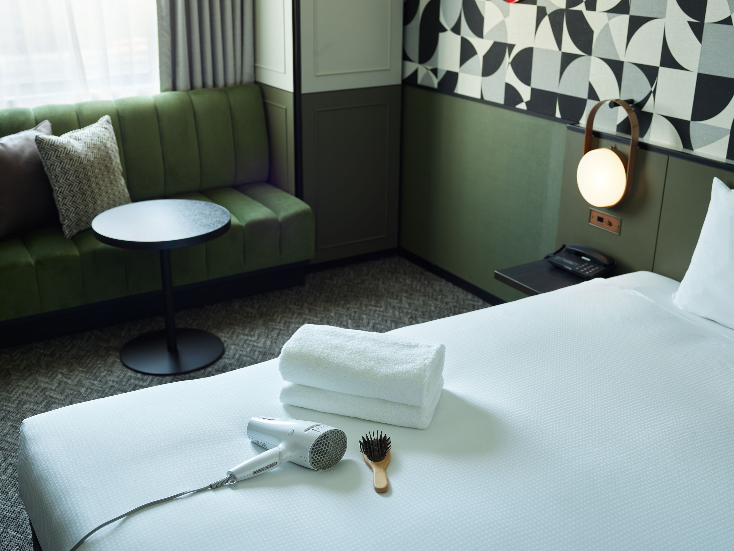 A hotel room with a bed, a hairdryer, a towel, a wooden brush, a green sofa, a small round table, and a wall-mounted lamp.