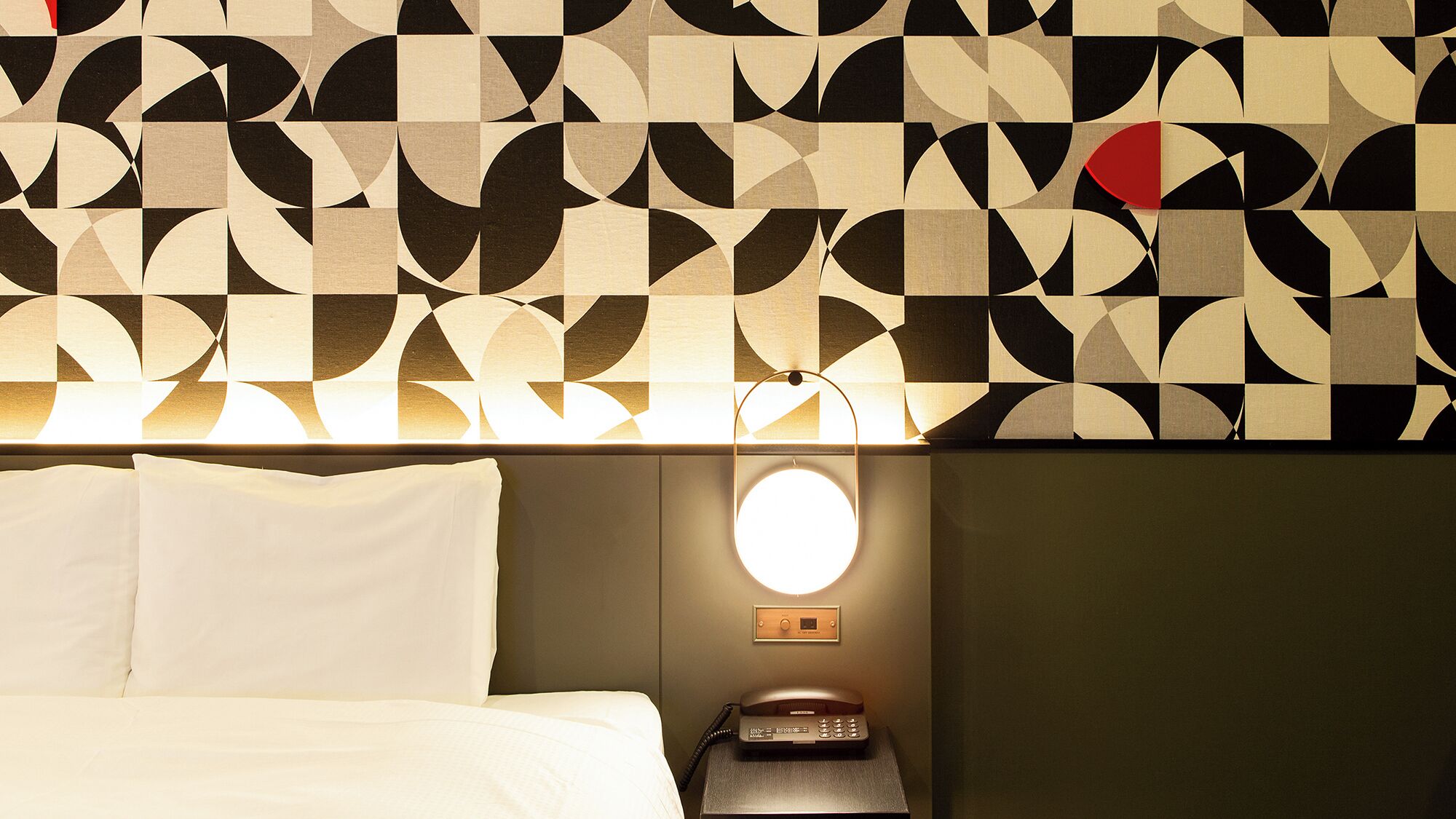 A hotel room with a geometric patterned wall, a bed with white linens, a bedside table with a lamp, and a phone.