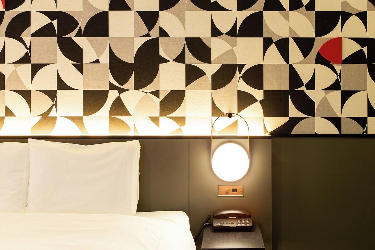 Habitación de hotel con pared de diseño geométrico, cama con ropa blanca, mesita de noche con lámpara y teléfono.