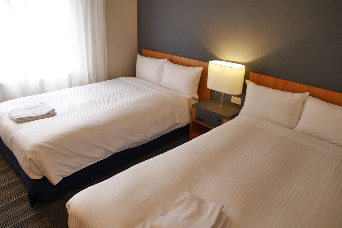Ein Hotelzimmer mit zwei Betten, jeweils mit weißer Bettwäsche und Kissen, einem Holzkopfteil, einem Nachttisch mit Lampe und einem Fenster mit leichtem Vorhang.