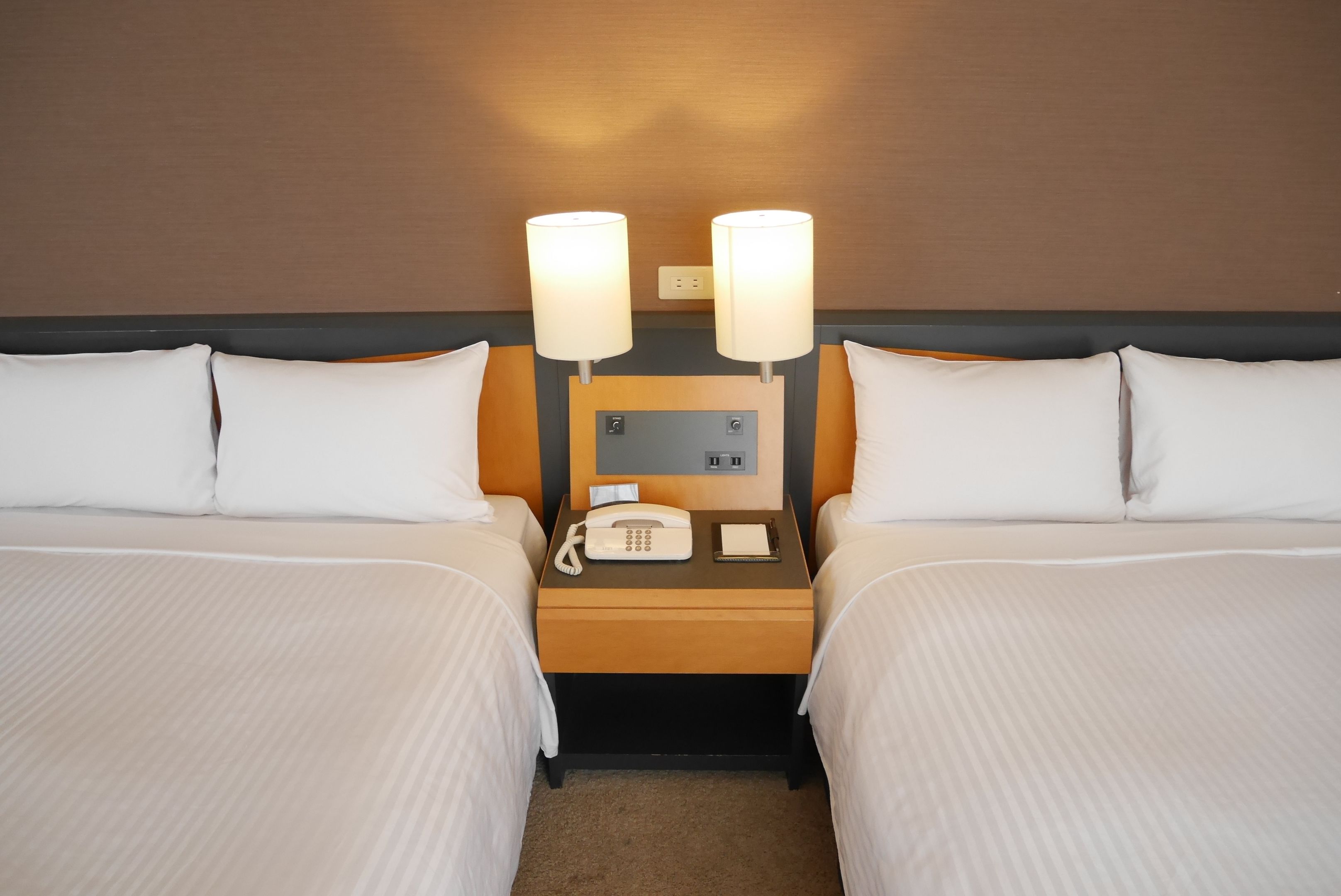 A hotel room with two beds, a nightstand with a phone and a notepad, and two wall-mounted lamps.