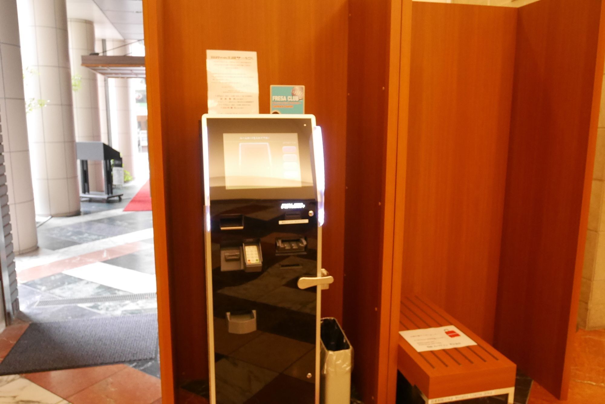A self-service kiosk in a hotel lobby with a trash bin and a small table with a sign.
