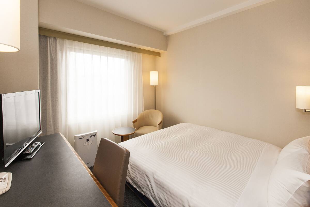 Ein Hotelzimmer mit einem Bett, einem Schreibtisch mit Computer, einem Stuhl, einer Lampe und einem Fenster mit Vorhängen.