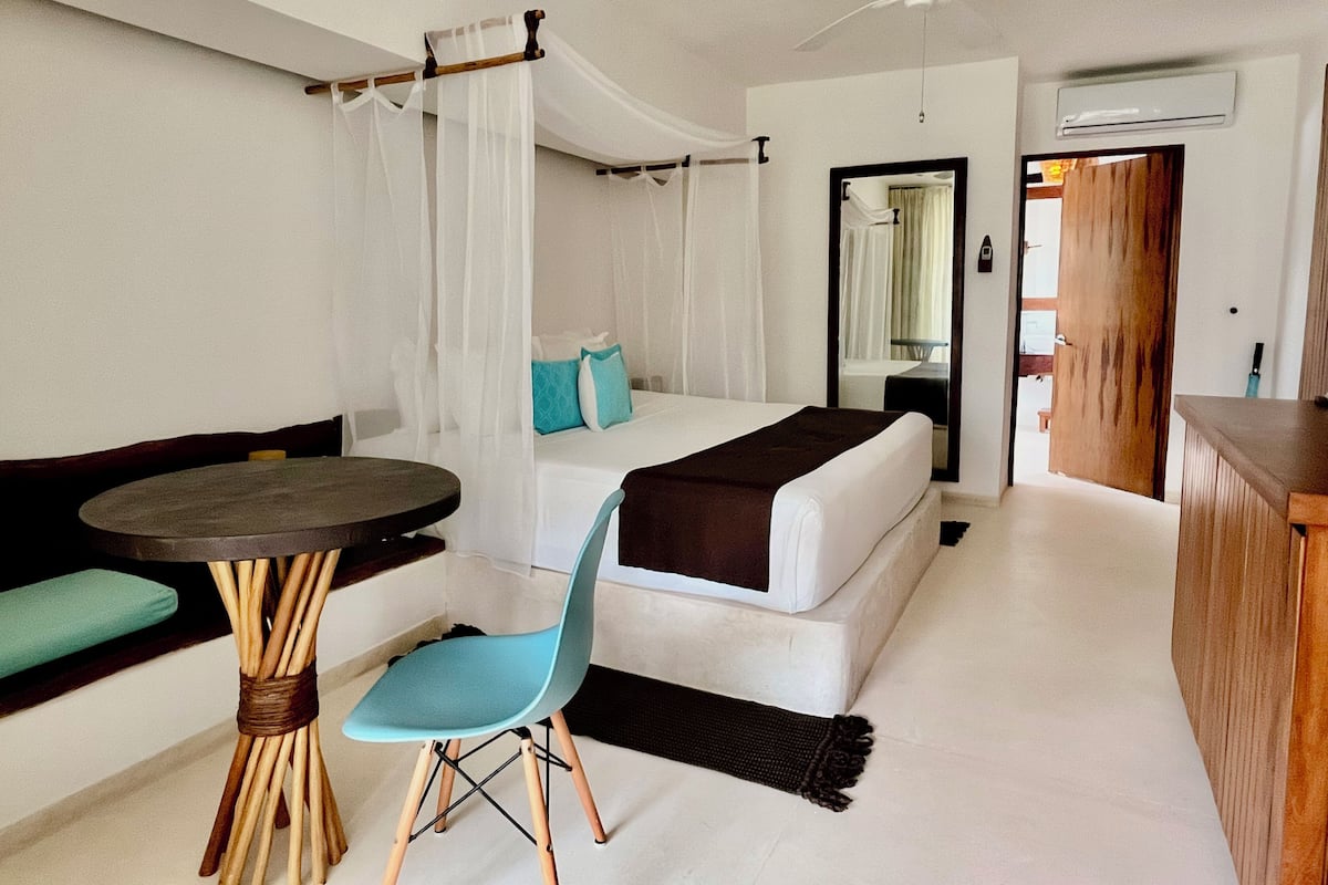 A hotel room with a bed, a chair, a small table, a ceiling fan, and a door leading to another room.