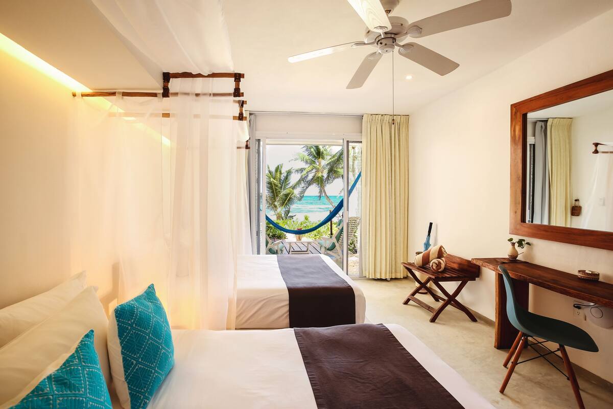 A hotel room with two beds, a desk, a chair, a ceiling fan, and a view of the ocean.