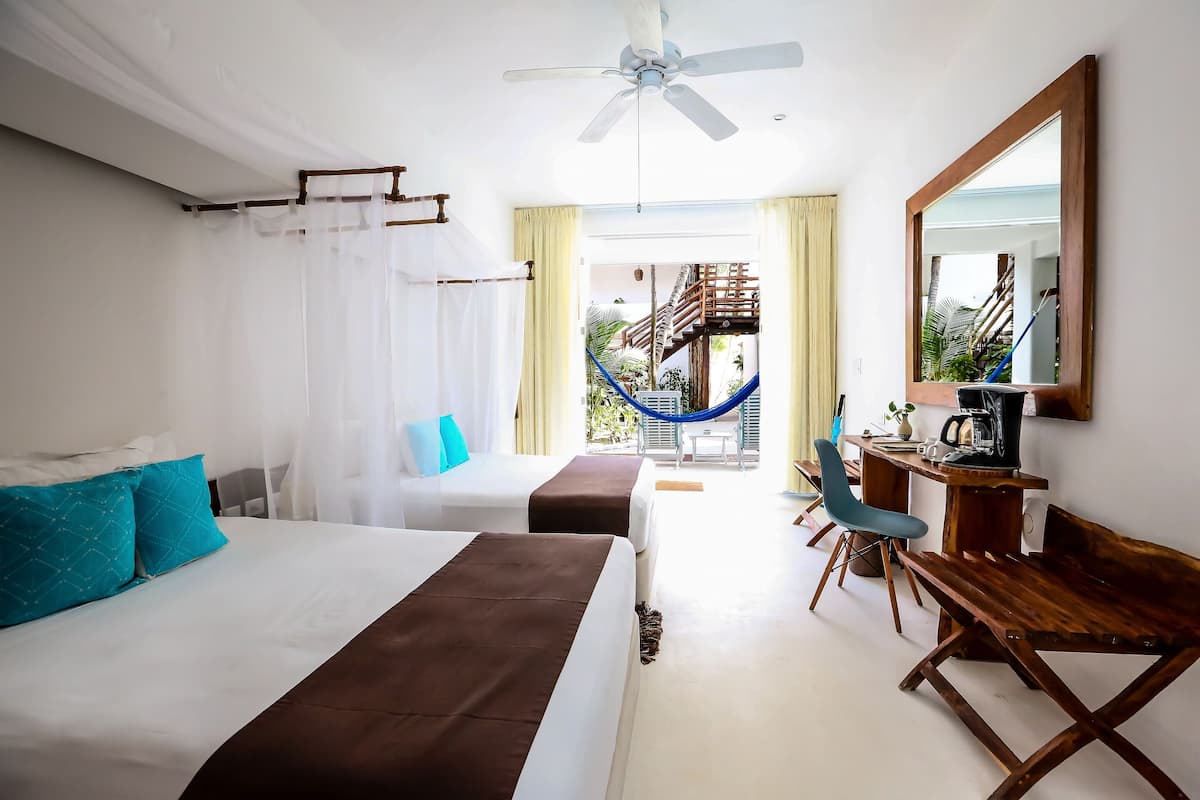 A hotel room with two beds, a desk with a chair, a ceiling fan, and a view of a staircase and outdoor area.