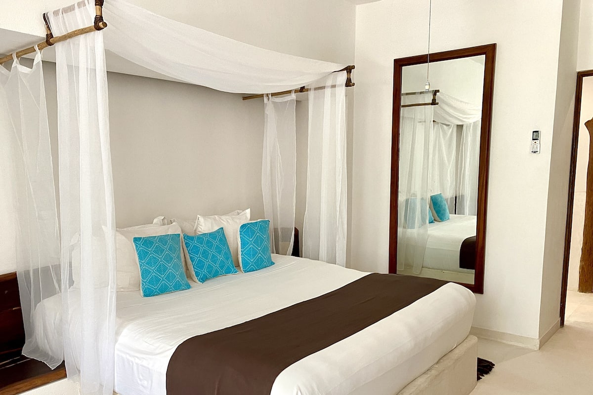 A neatly made bed with white and brown linens, a canopy, and blue patterned pillows.