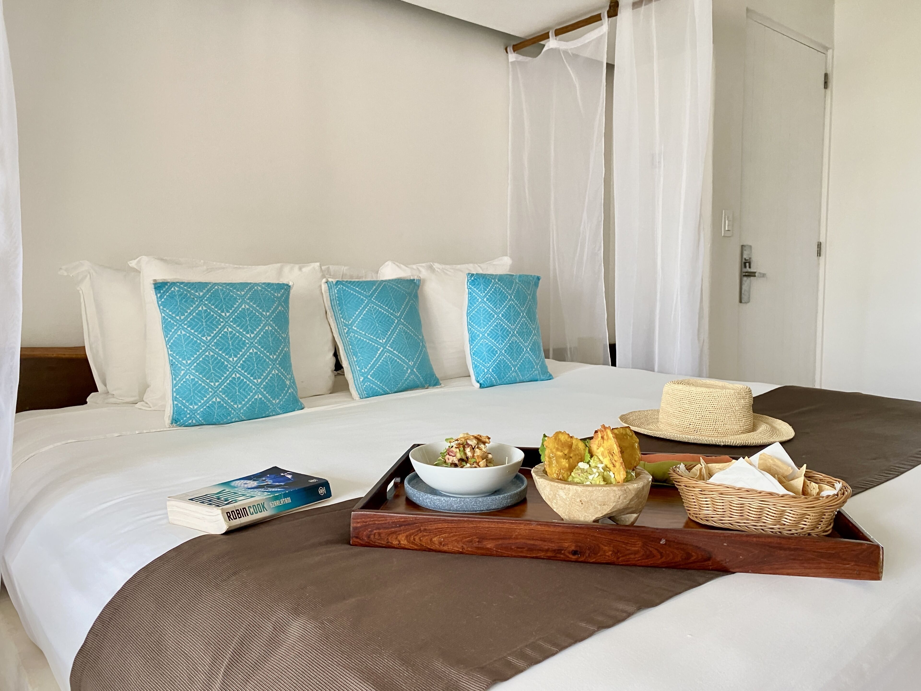 Una cama bien tendida con una bandeja de desayuno, que incluye pan, un bol de fruta y un libro titulado 