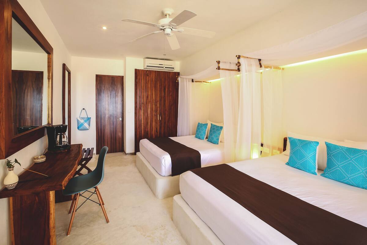 A hotel room with a wooden desk, two beds, a ceiling fan, and a bathroom with a shower curtain.