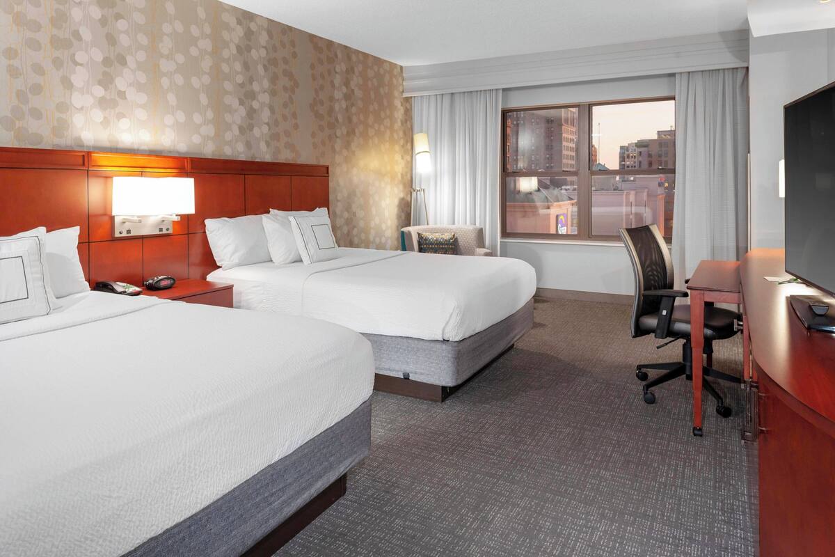 A hotel room with two beds, a desk, a chair, a TV, and a window with a city view.