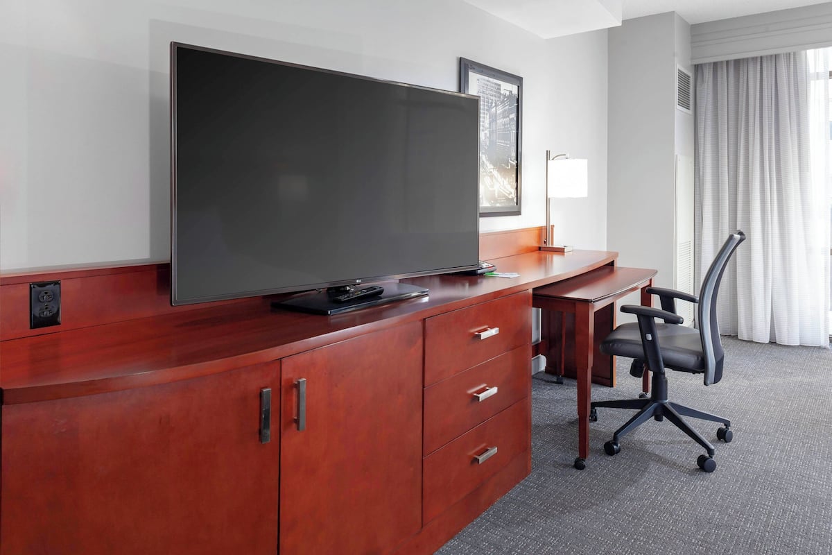 A hotel room with a large flat-screen TV on a wooden desk, a chair, and a framed picture on the wall.
