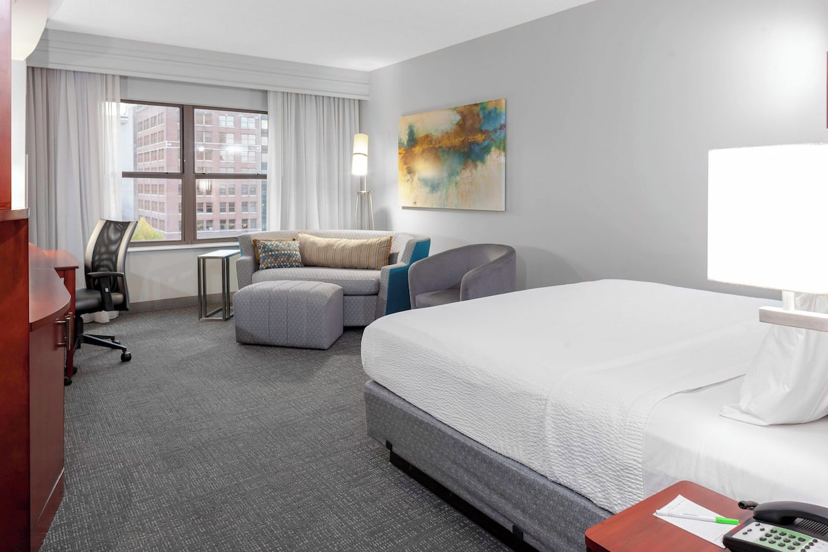 A hotel room with a large bed, a desk with a lamp, a chair, a small table, and a window with curtains.