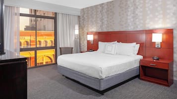 A hotel room with a large bed, a nightstand, a desk, and a chair. There is a window with a view of a building.
