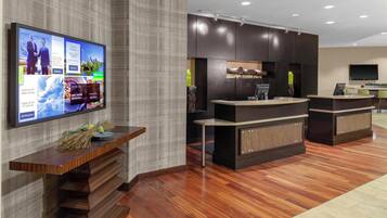 Una zona de recepción con un mostrador de madera, un televisor de pantalla plana mostrando varios anuncios, y un diseño interior moderno con paneles de madera oscura y suelo de baldosas.