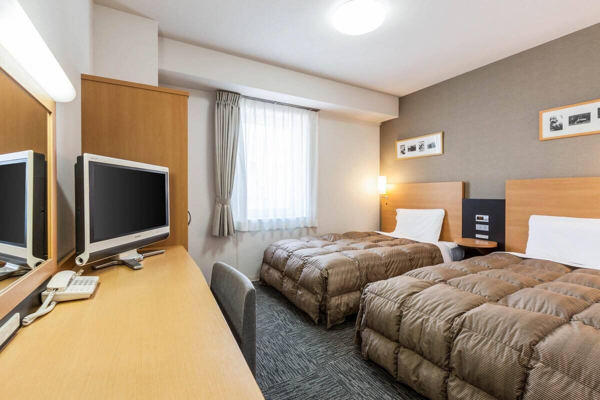 A hotel room with two beds, a desk with a computer and phone, a window with curtains, and framed pictures on the wall.