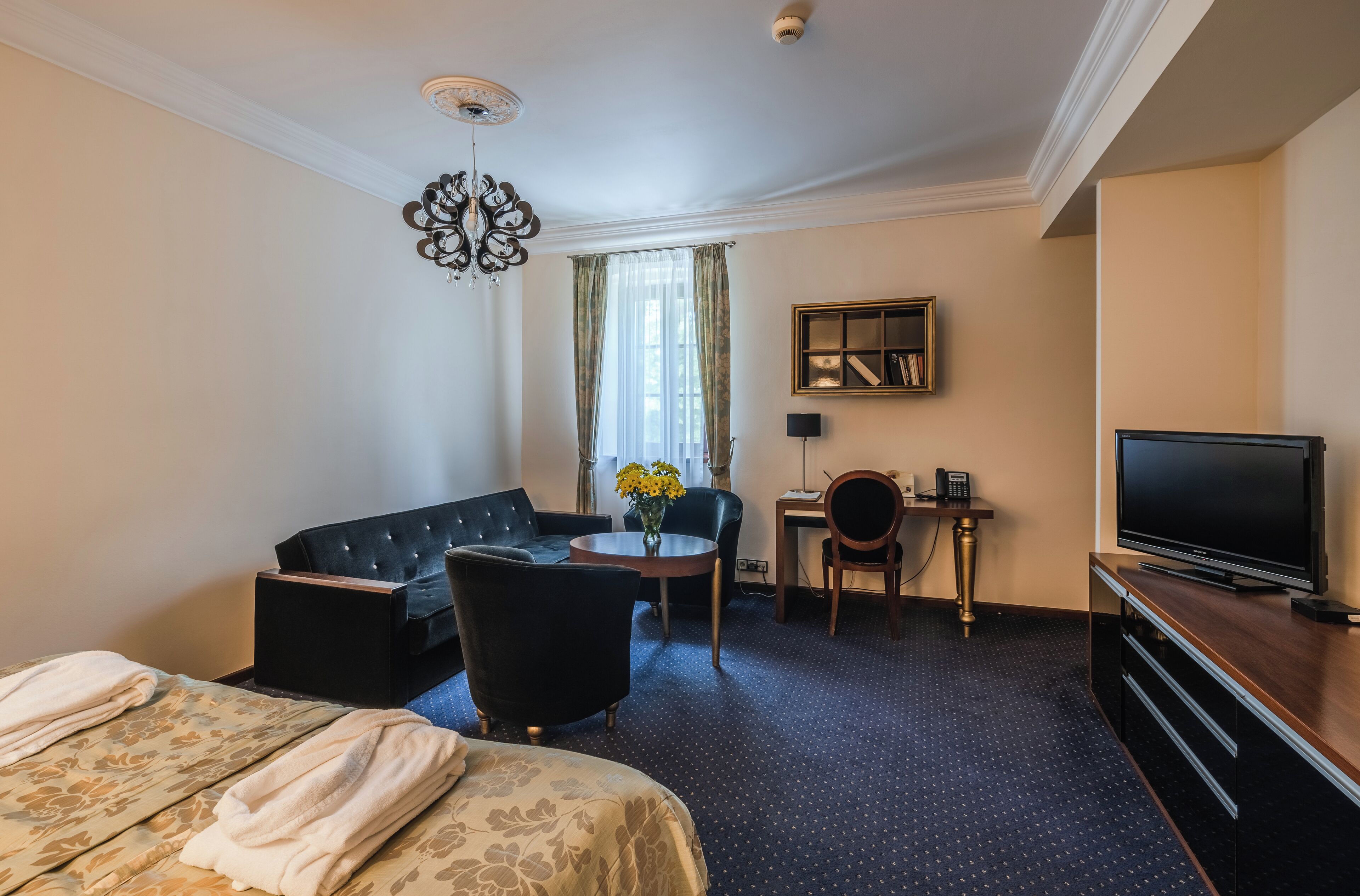A hotel room with a bed, a sofa, a desk, a chair, a television, and a chandelier.