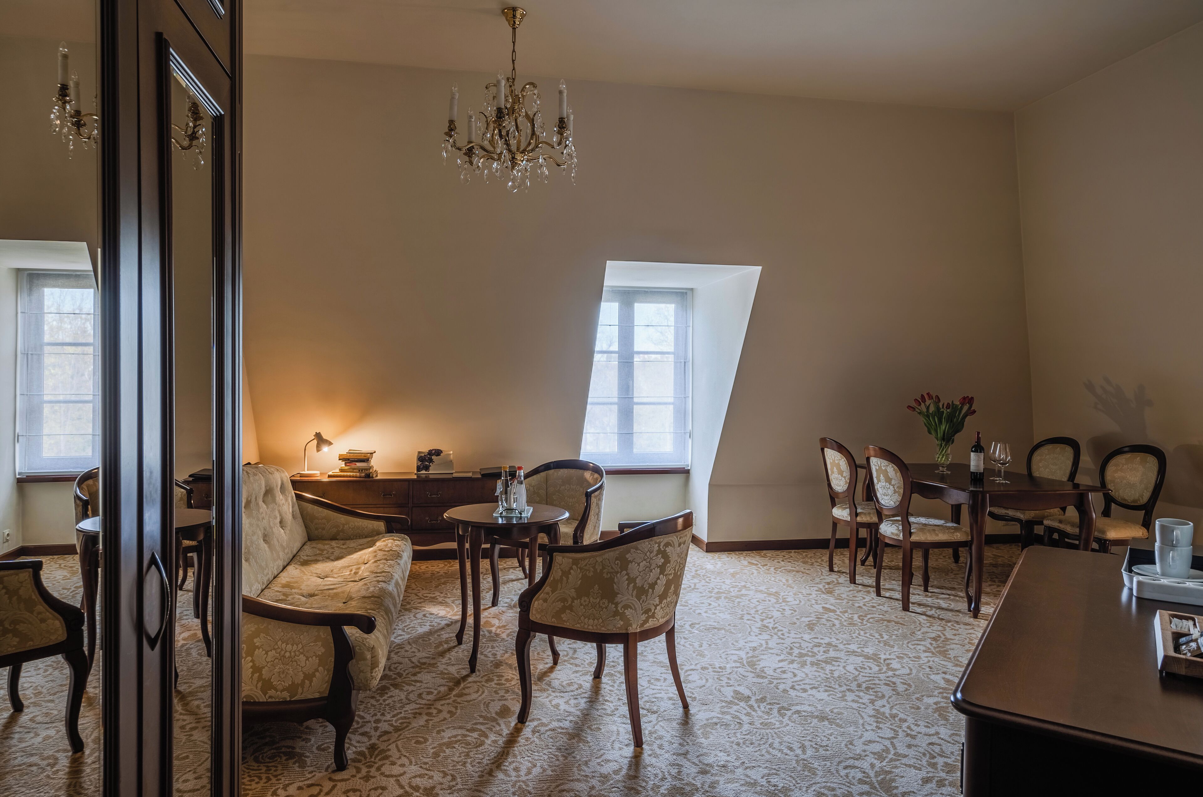 Una habitación amplia con techo alto, un candelabro, una mesa de comedor con sillas, un sofá y un ventanal que proporciona luz natural.