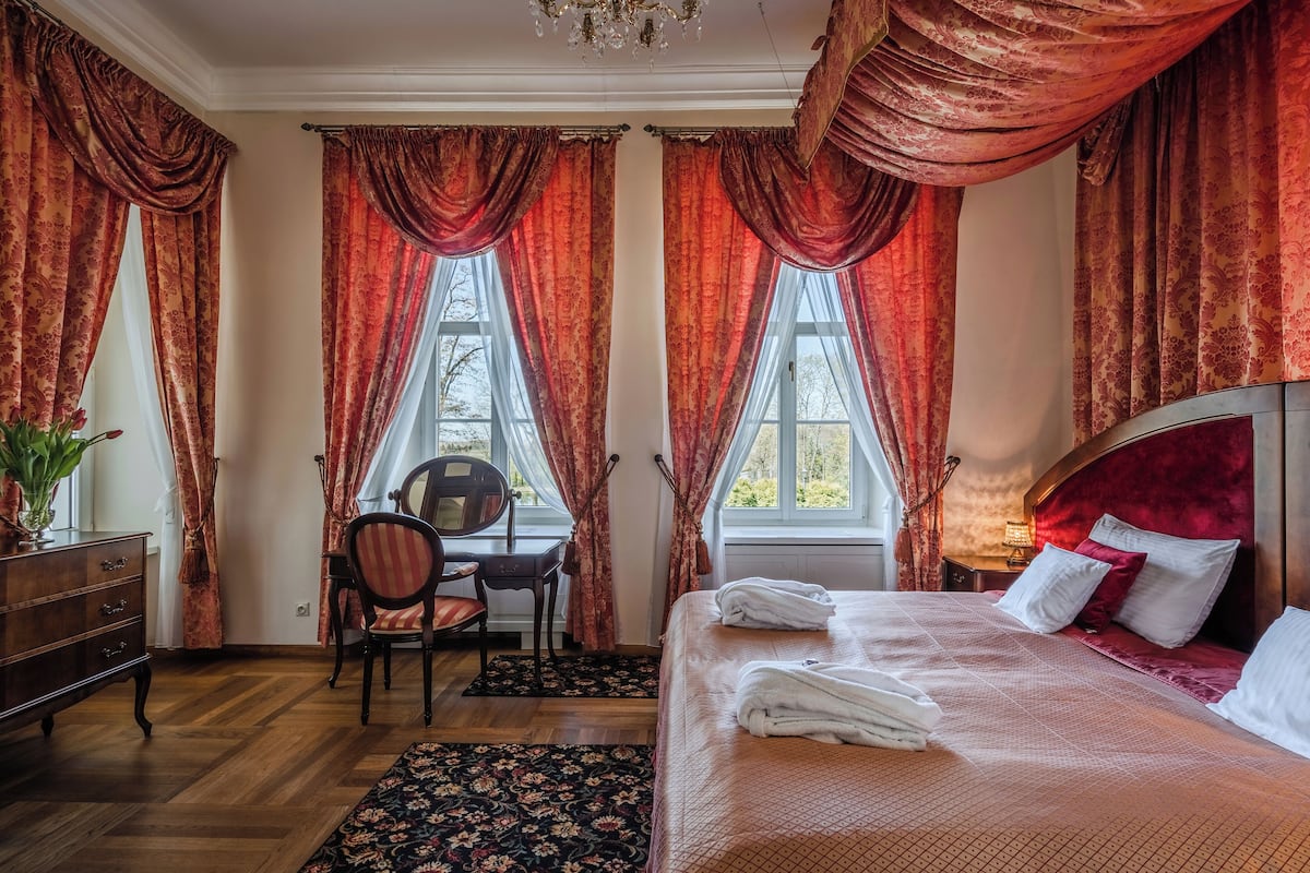 Une chambre à coucher avec un grand lit, un bureau avec une chaise et deux fenêtres ornées de rideaux rouges.