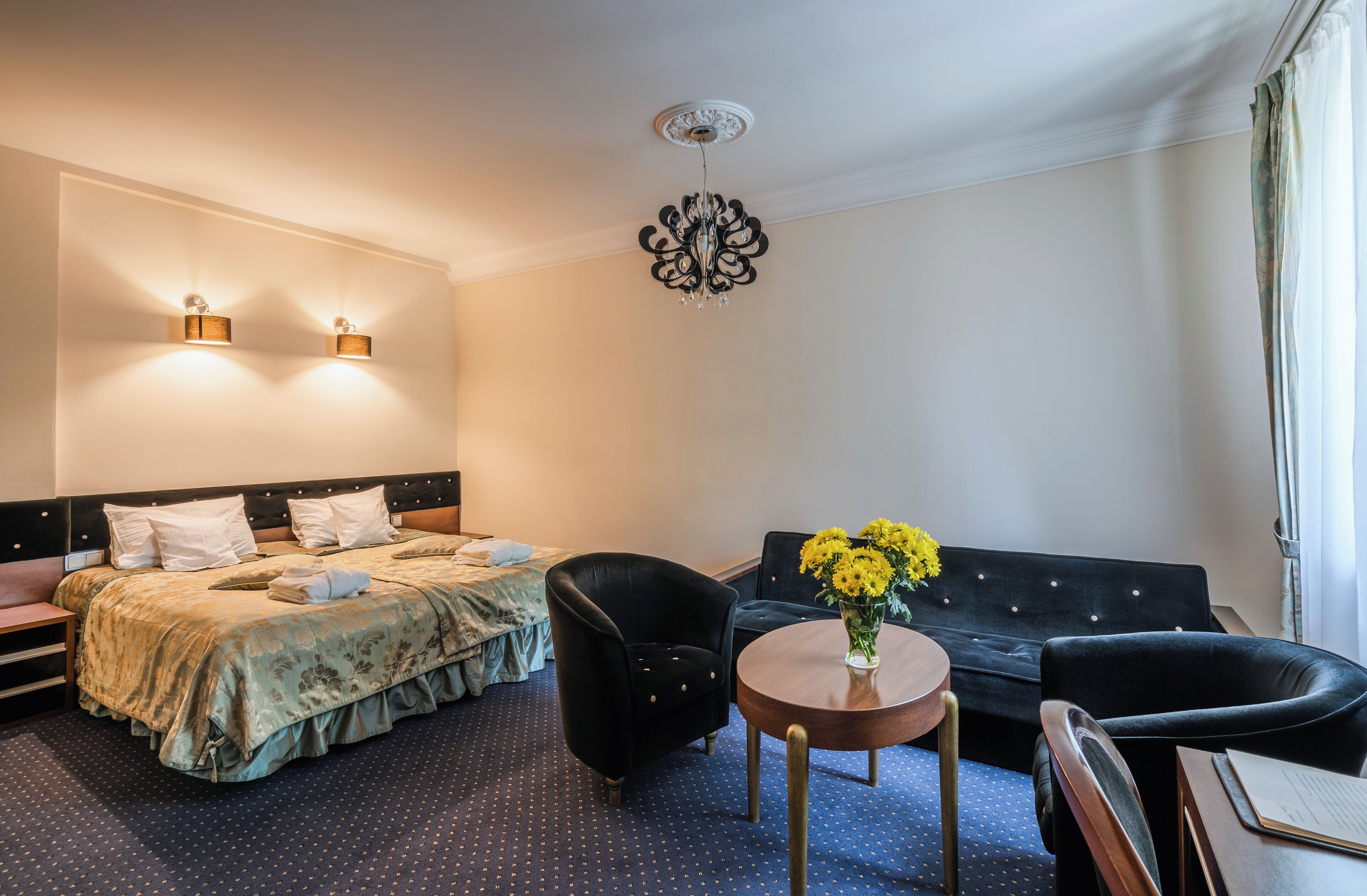 Habitación de hotel con cama, dos sillones, un sofá, una mesita con un jarrón de flores y una lámpara en el techo.