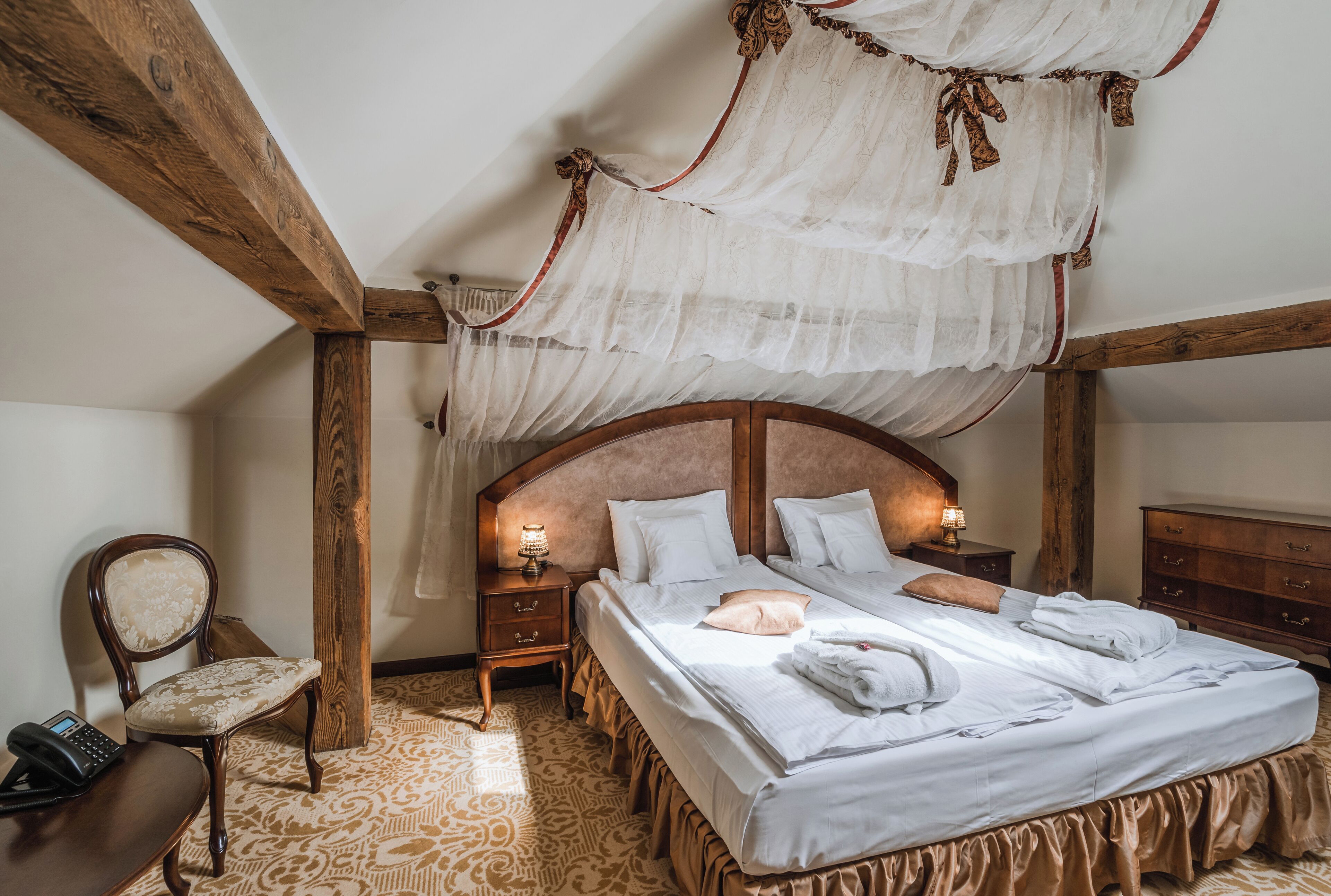 A bedroom with a canopy bed, wooden headboard, bedside tables with lamps, a chair, and patterned carpet.