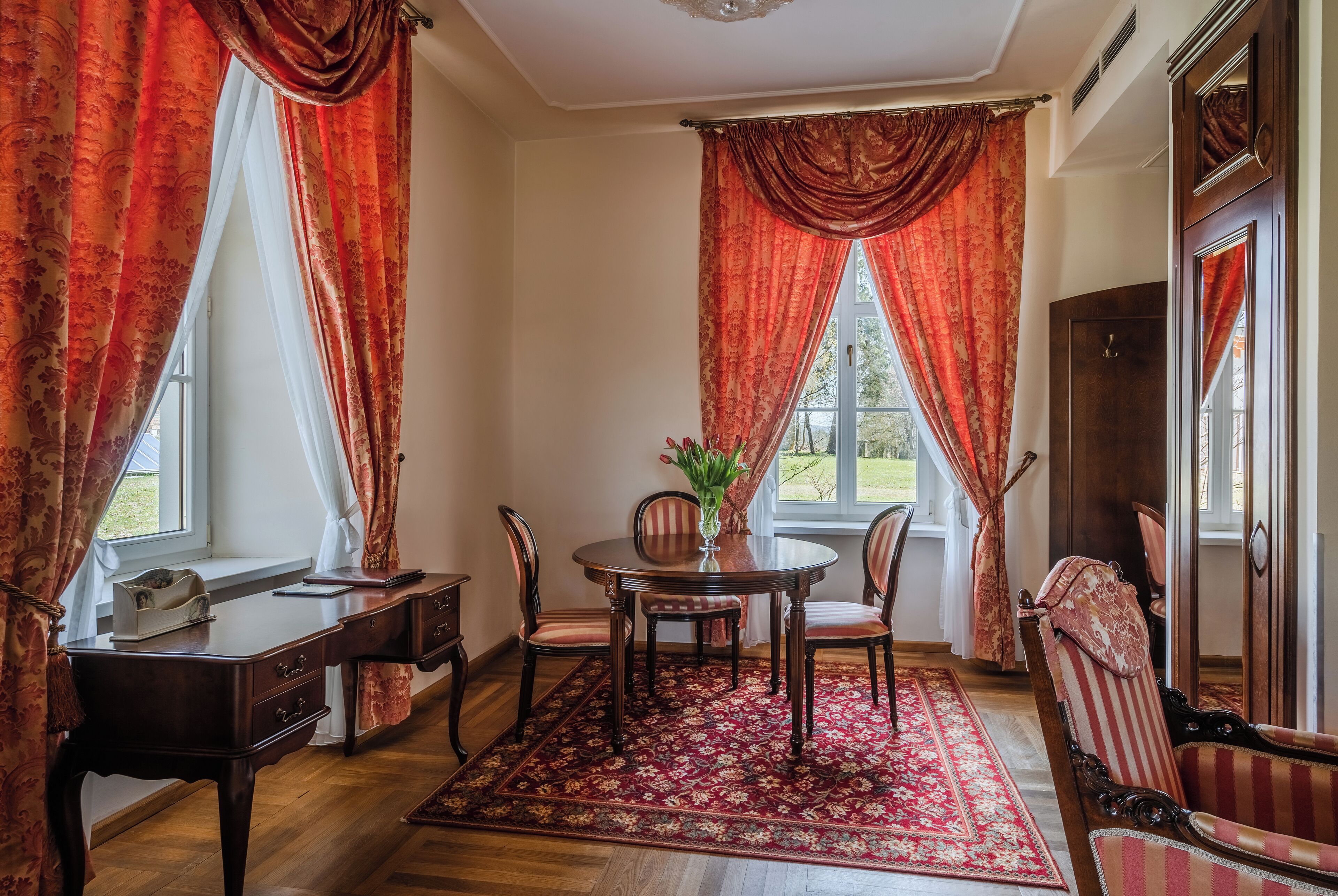 A room with a wooden desk, a round table with chairs, a red patterned rug, and a large window with red curtains.