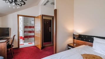 A hotel room with a bed, a desk with a chair, a TV, a wardrobe, and a shower area.
