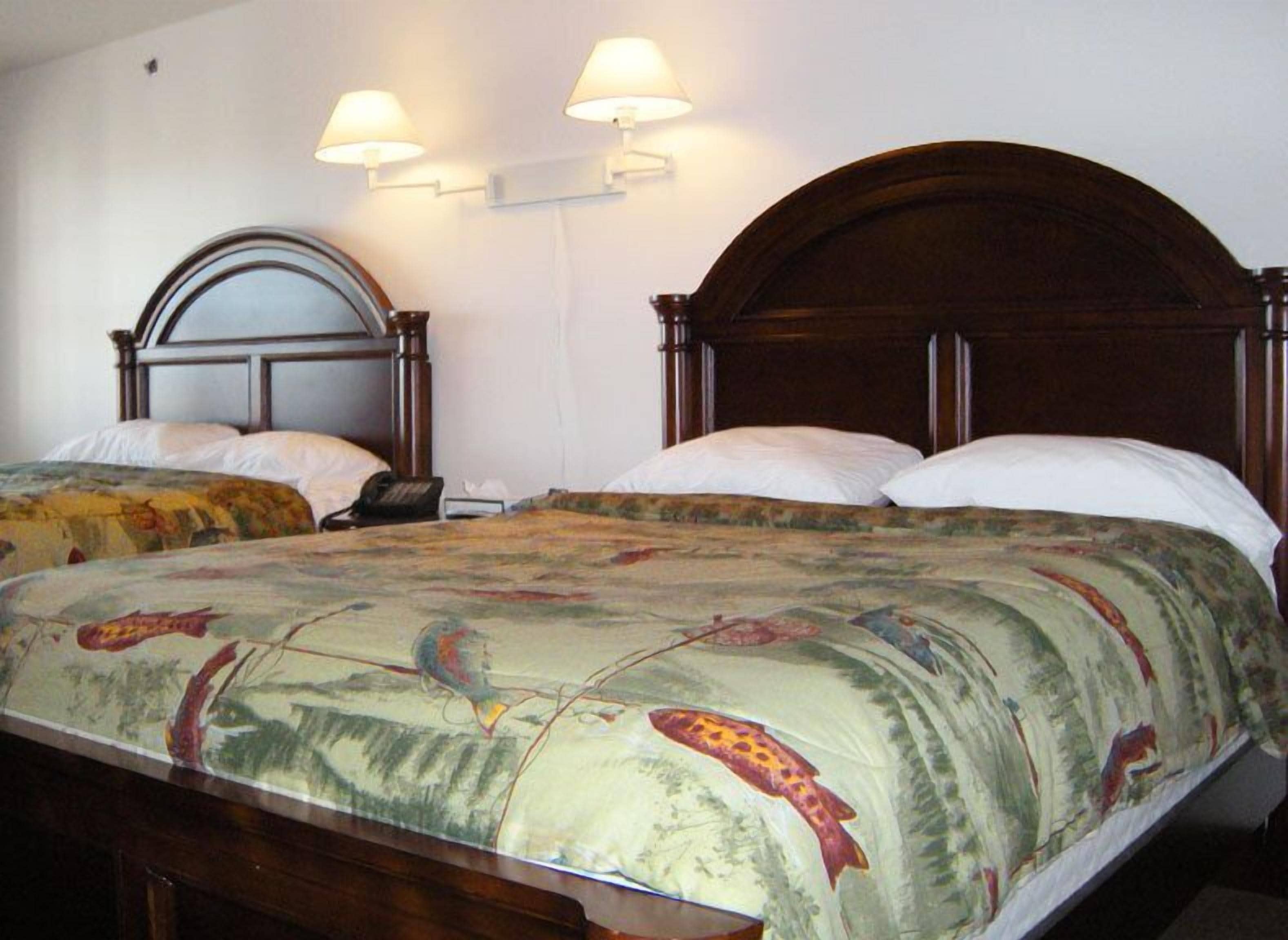 A hotel room with two wooden beds, each with a headboard and white bedding. There are wall-mounted lamps on either side of the beds.
