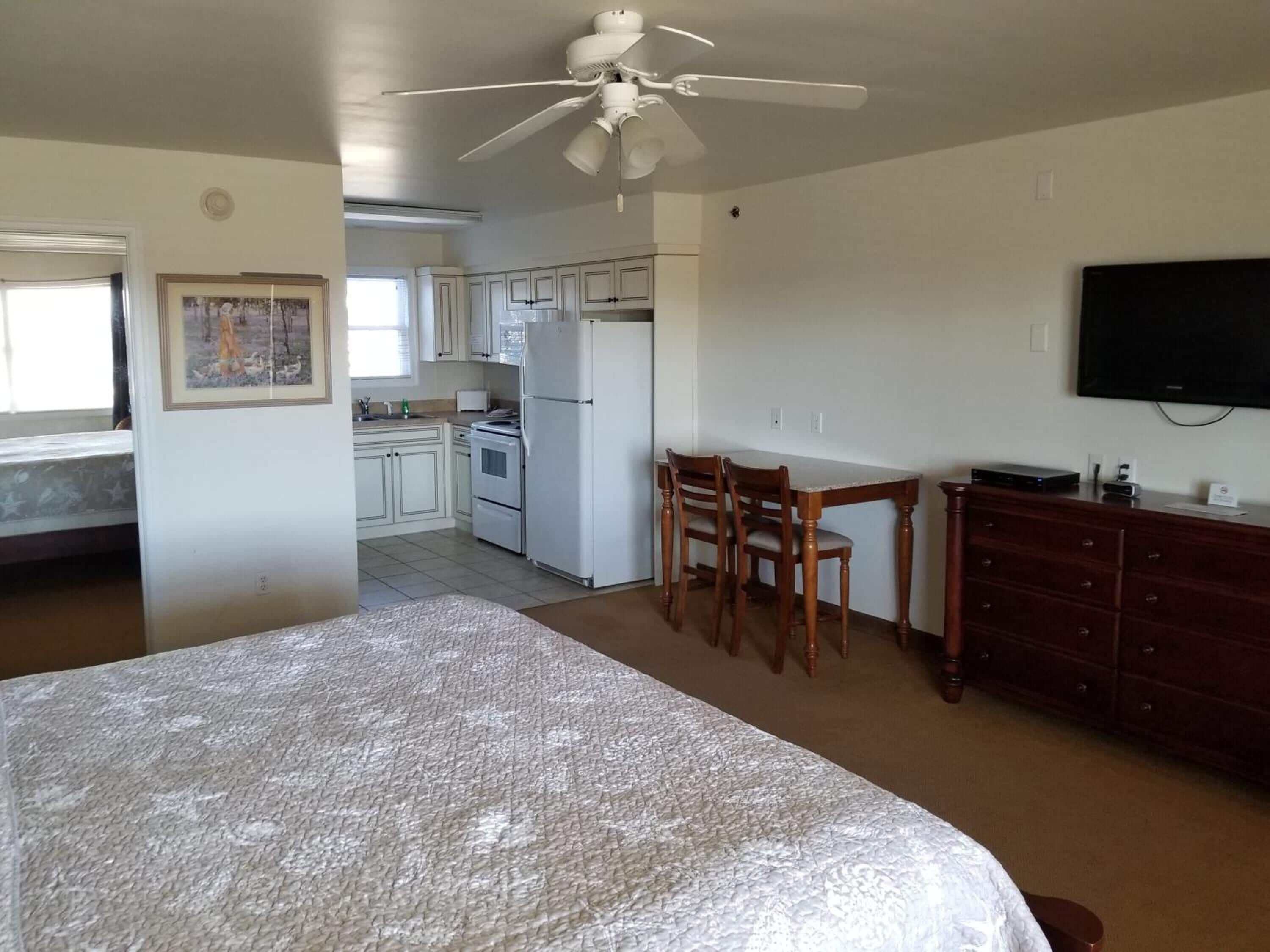 Ein Schlafzimmer mit Bett, Deckenventilator, Esstisch mit Stühlen, Küchenzeile mit Schränken und Geräten sowie einem an der Wand montierten Fernseher.