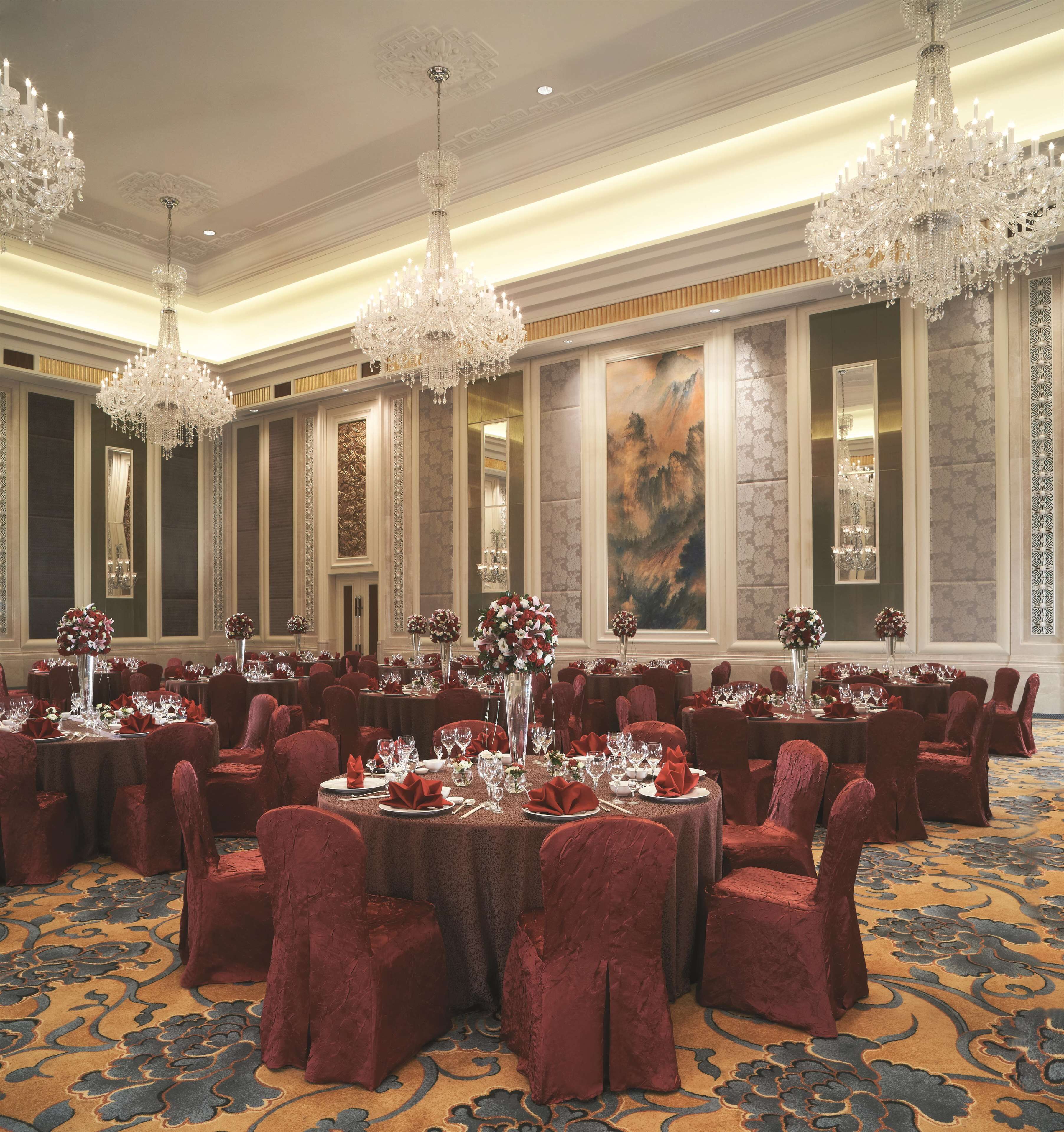 Een feestzaal met ronde tafels, rode fluweelstoelen, kroonluchters en bloemstukken als decoratie.