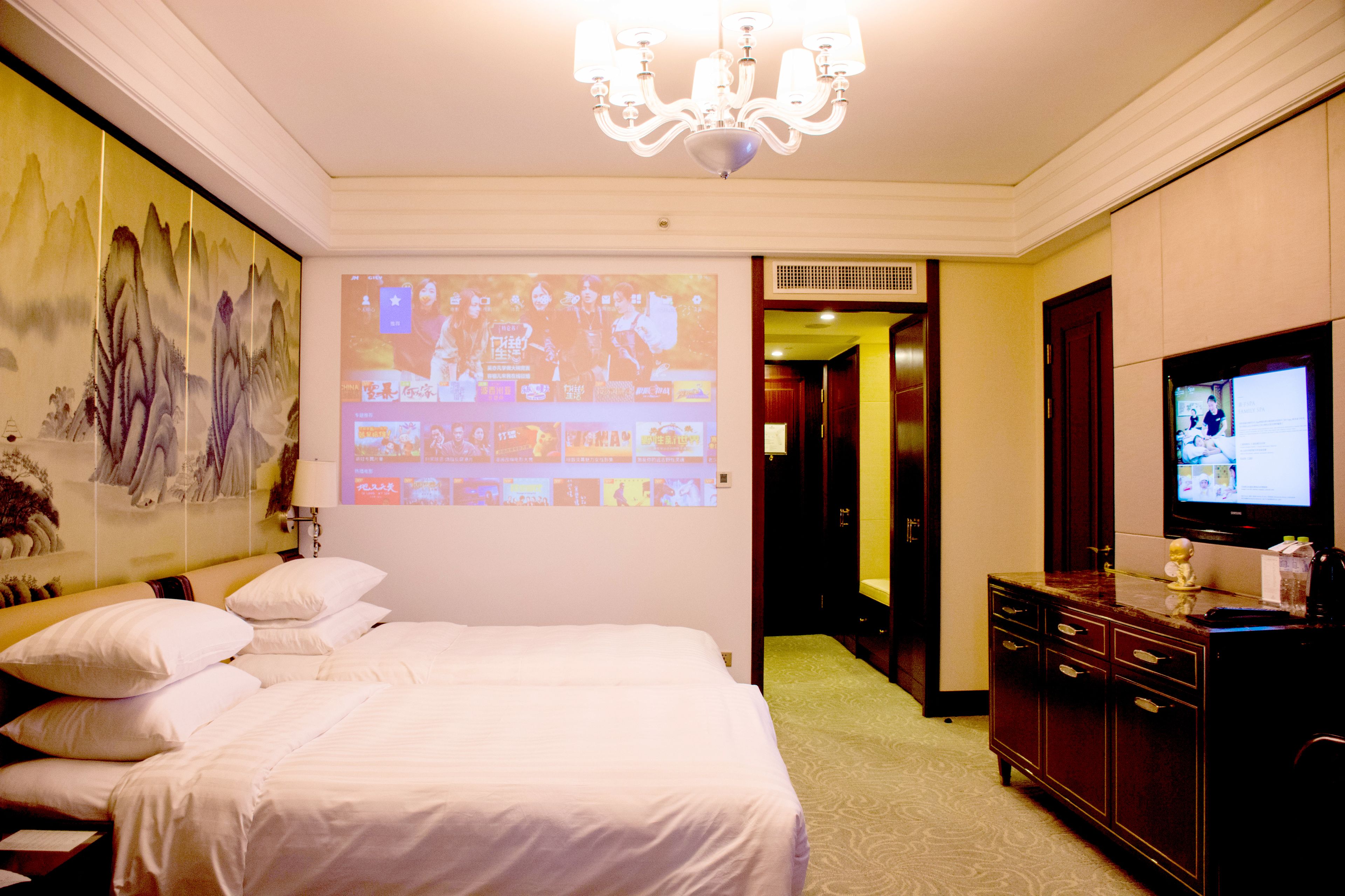 Una habitación de hotel con una cama grande, televisión, cómoda y una pantalla de proyección mostrando un evento deportivo.