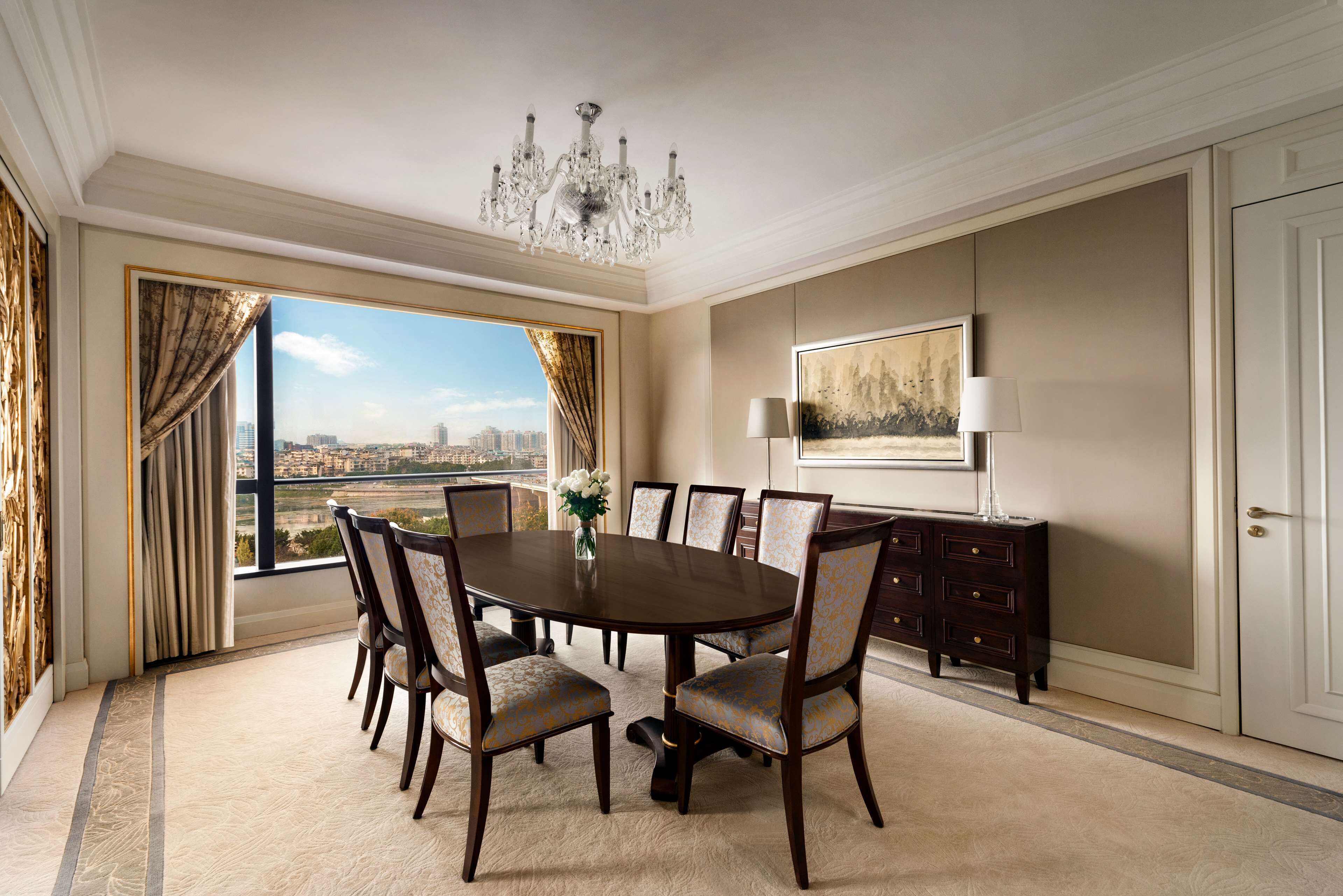 A spacious dining room with a large round table, six chairs, a chandelier, a city view, a framed artwork, and a cabinet.