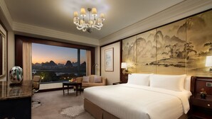A hotel room with a large bed, a desk, a chair, a view of the cityscape at sunset, and a large painting on the wall.