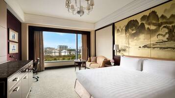 A hotel room with a large bed, a desk, a chair, a view of the city, and a painting on the wall.