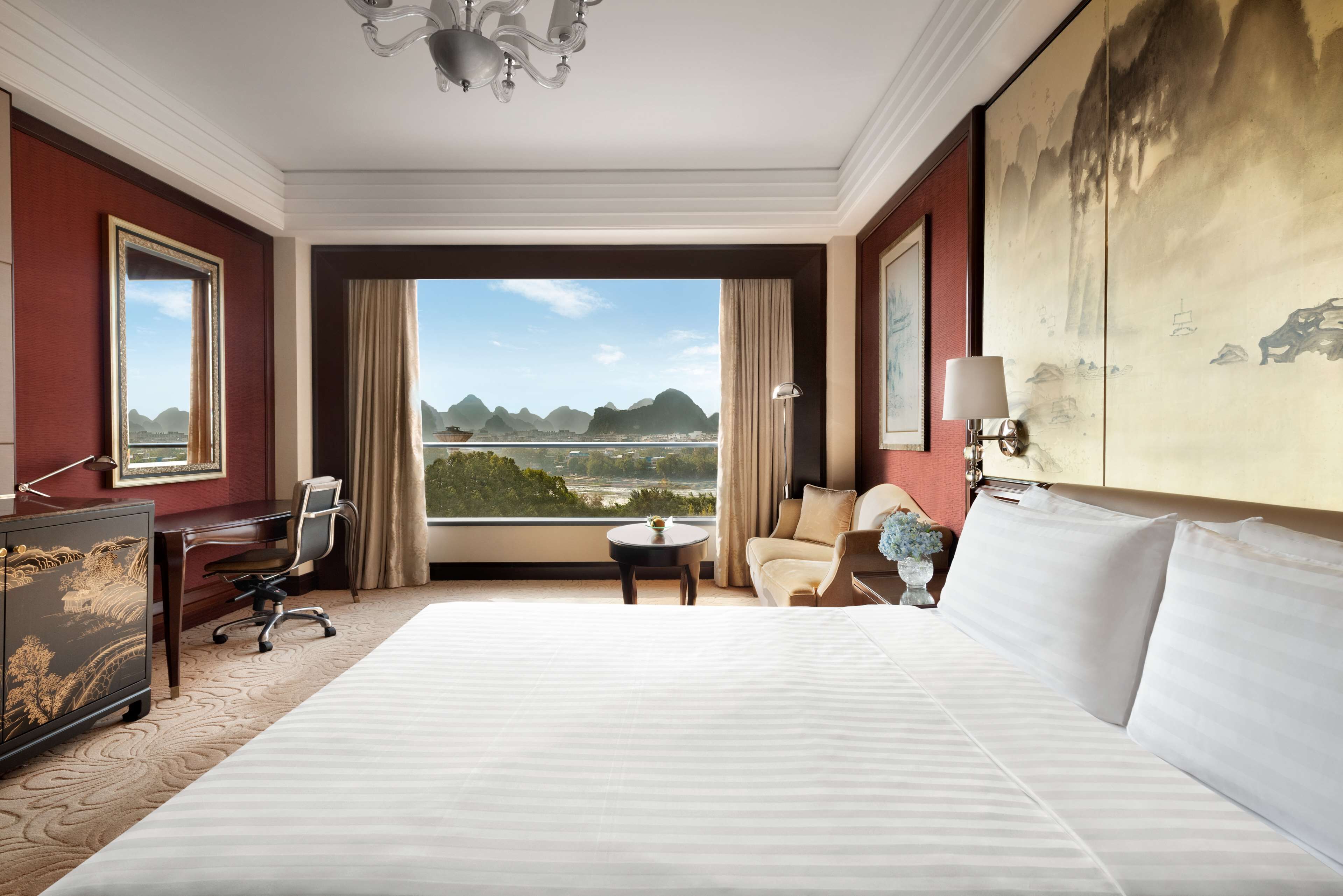 Habitación de hotel con una cama grande, un escritorio, una silla, una mesita y vistas a las montañas desde la ventana.