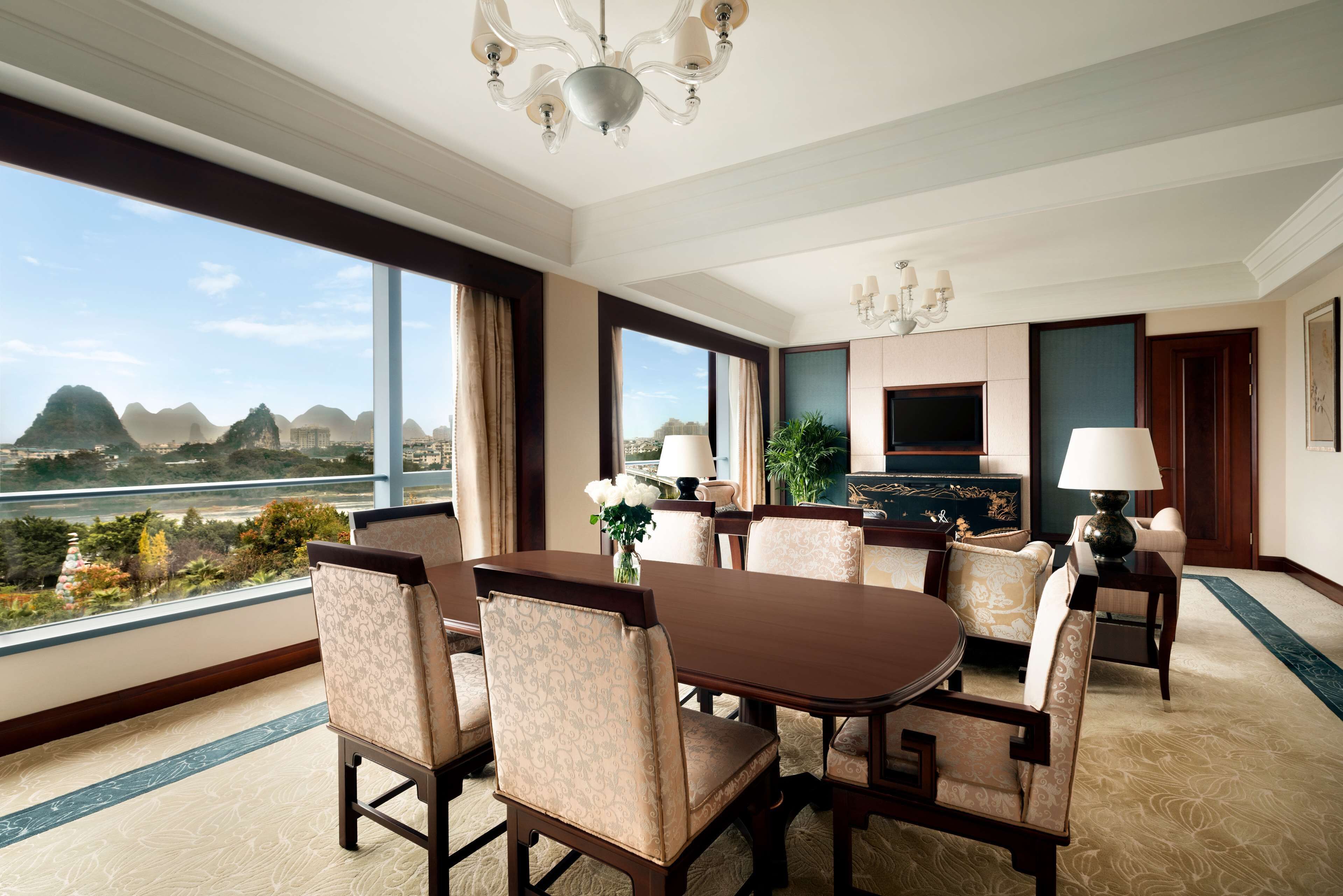 A hotel room with a dining area, large windows with a view of mountains, a chandelier, and a flat-screen TV.