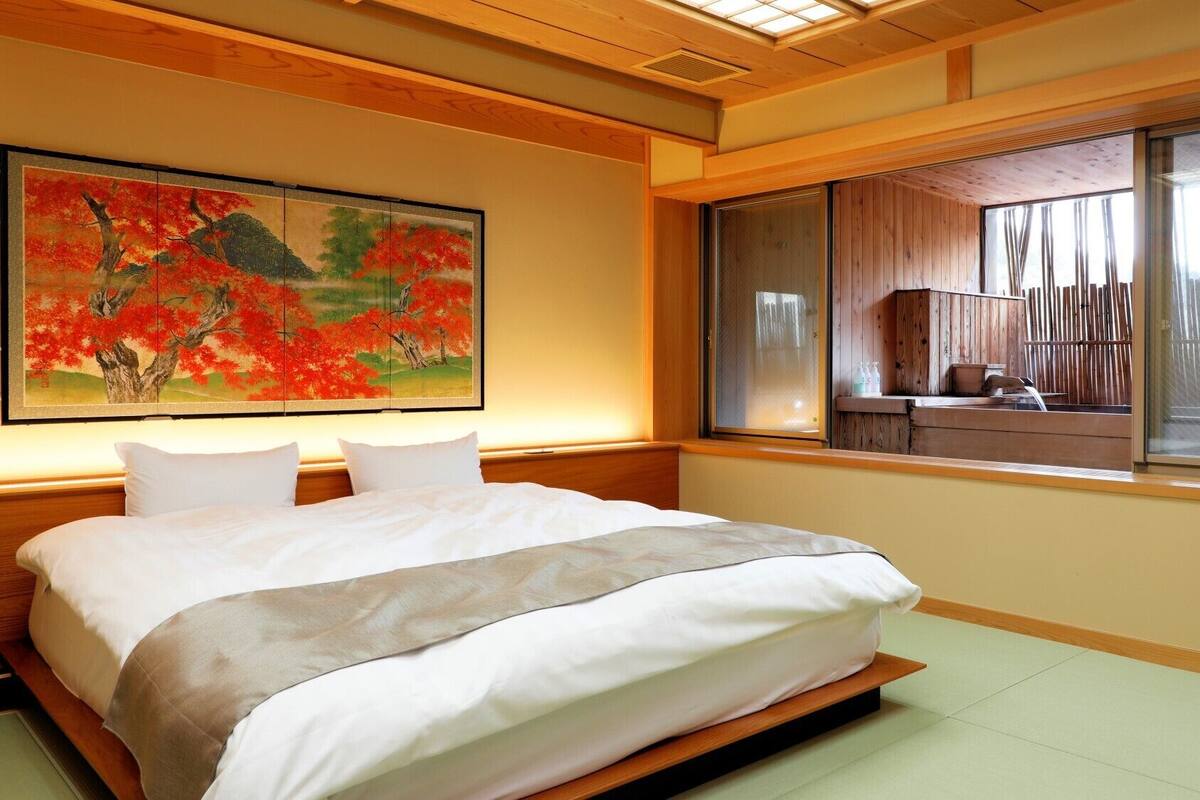 Ein Hotelzimmer mit einem großen Bett, einem Gemälde von roten Bäumen, einem Fenster mit Blick auf einen Holzzaun und einer kleinen Holzkabine.