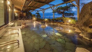 A stone hot spring bath with wooden beams overhead, surrounded by rocks and trees, with a view of the sea. - Inatori-Ginsuiso (Higashiizu)