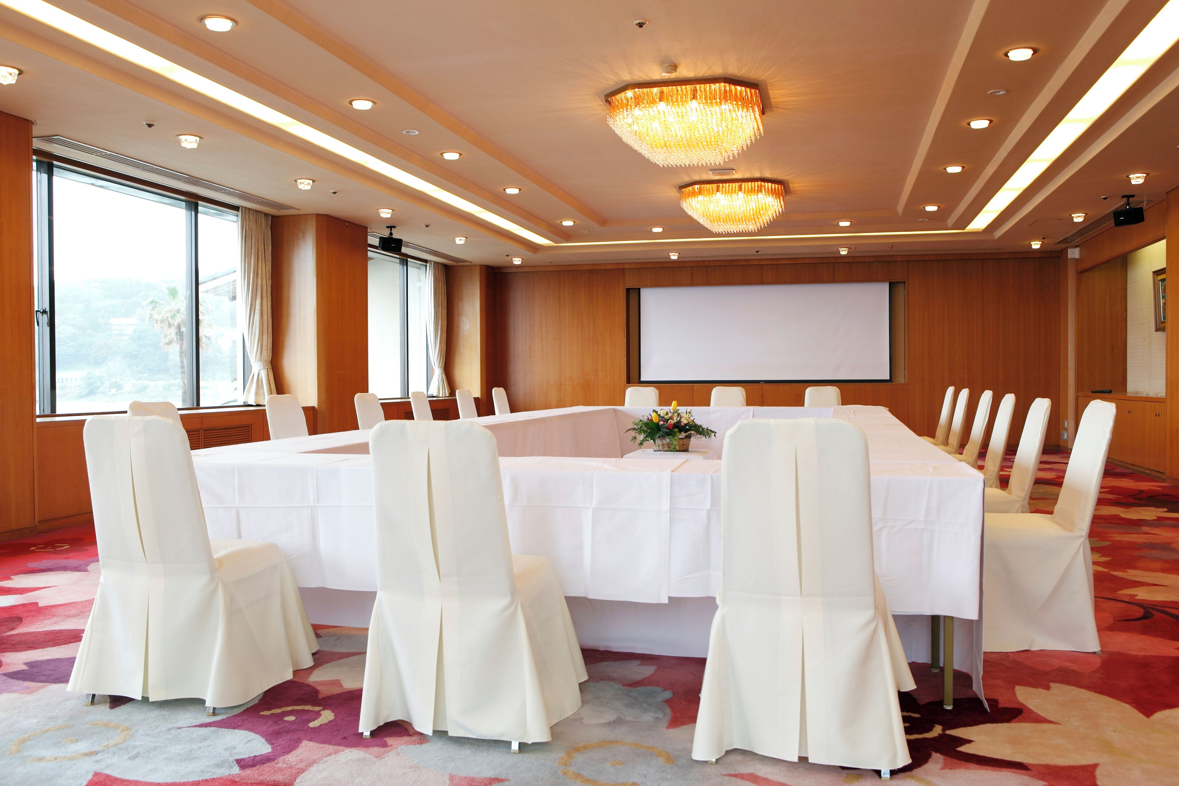 Une salle de conférence avec une longue table, des chaises blanches, un lustre, un écran de projection et de grandes fenêtres.