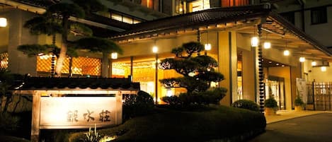 Un edificio japonés tradicional de varios pisos, con un acceso cubierto y jardines bien cuidados que incluyen bonsáis.