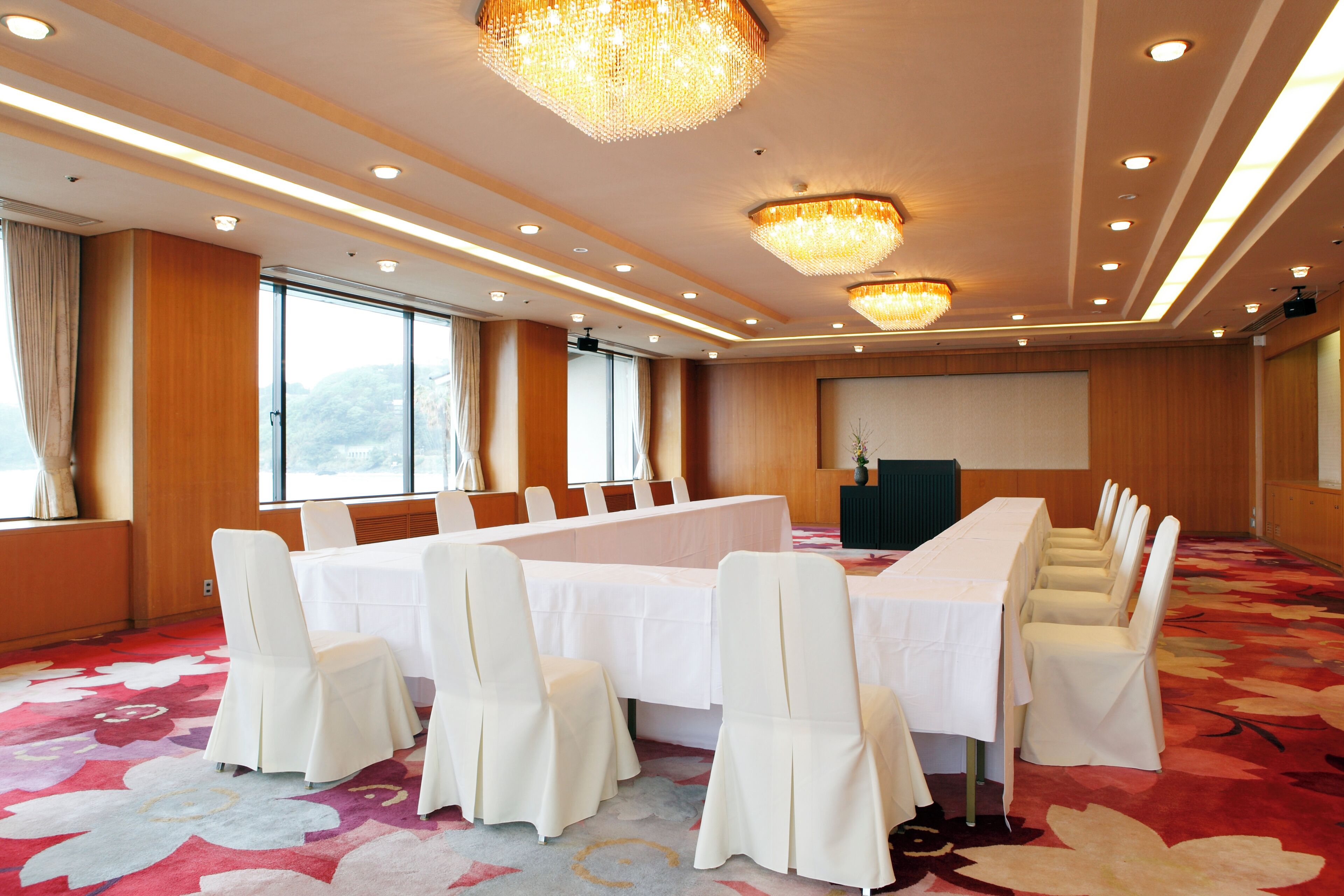 A conference room with a long table, white chairs, chandeliers, and large windows.