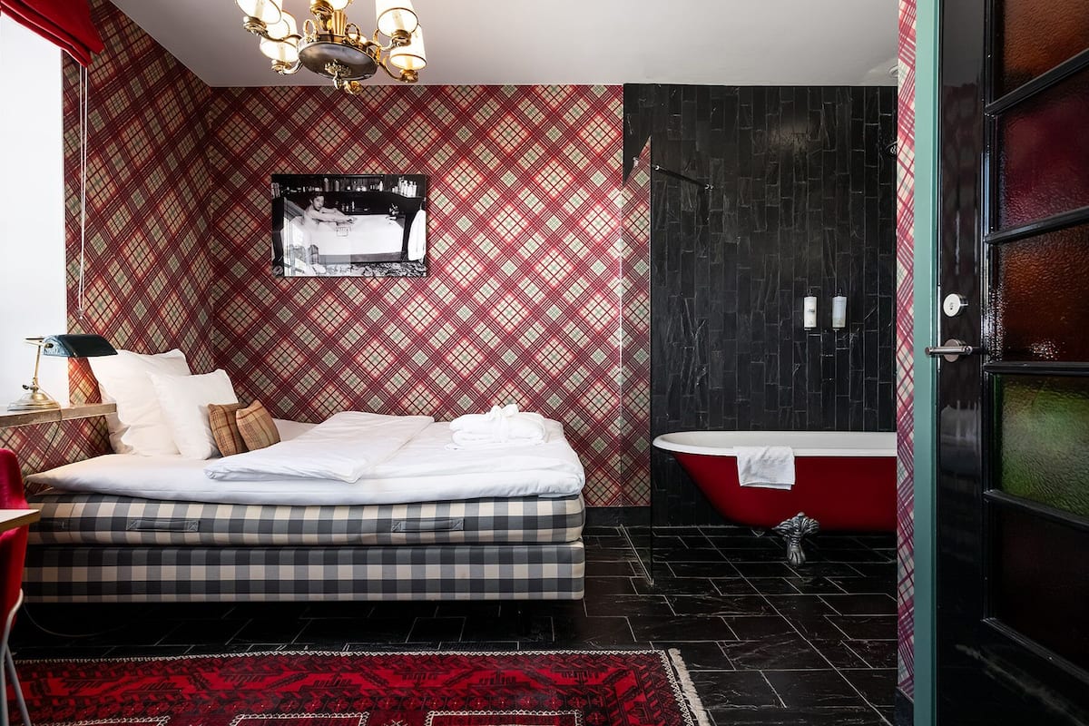 A bedroom with a bed, a red and black checkered wall, a black bathtub, and a red door.