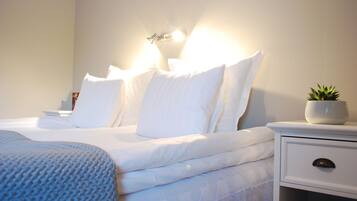 A neatly made bed with white linens and pillows, a bedside table with a plant, and a wall-mounted light fixture.